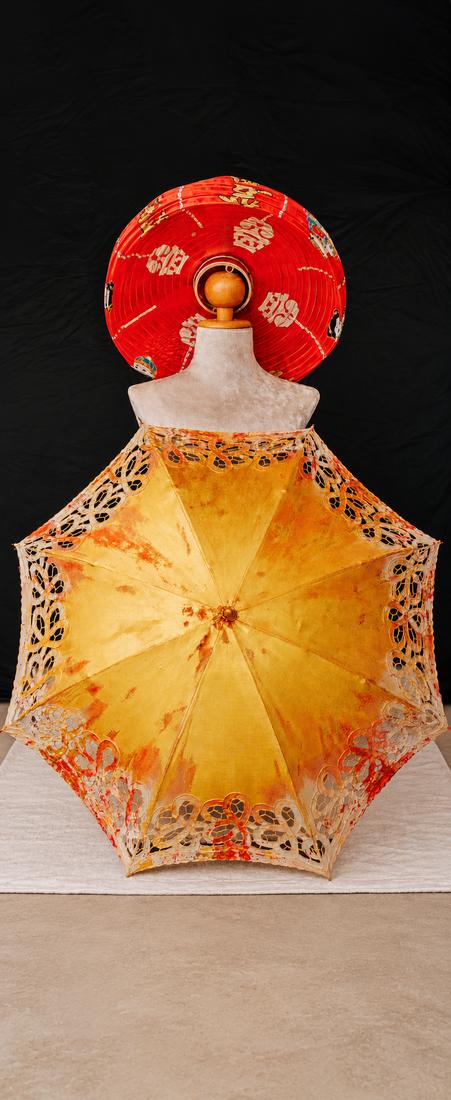 Open parasol of gold silk with cut lace edges and red paint, in front of a dress form wearing a round red hat with gold symbols, on black.
