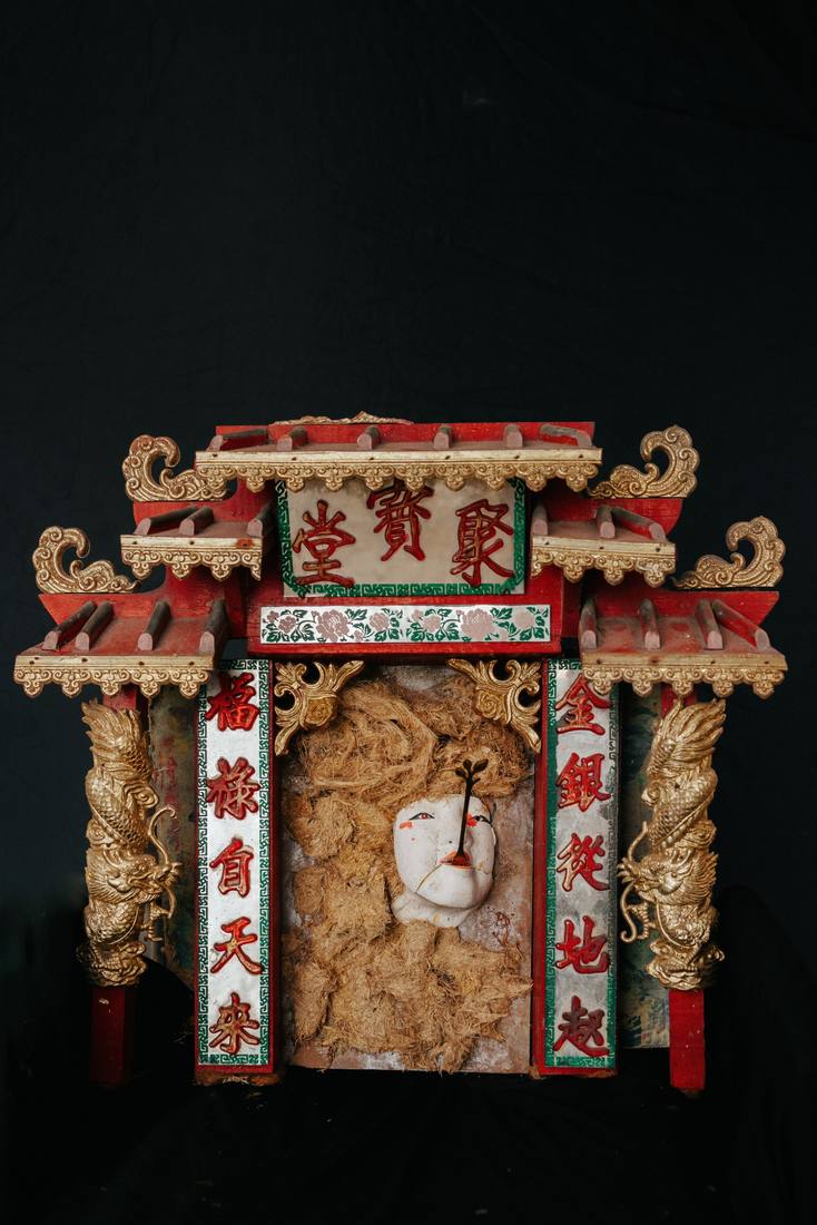 Small red and gold shrine with Chinese lettering on its pillars, a cracked white face in fibre inside, on black.
