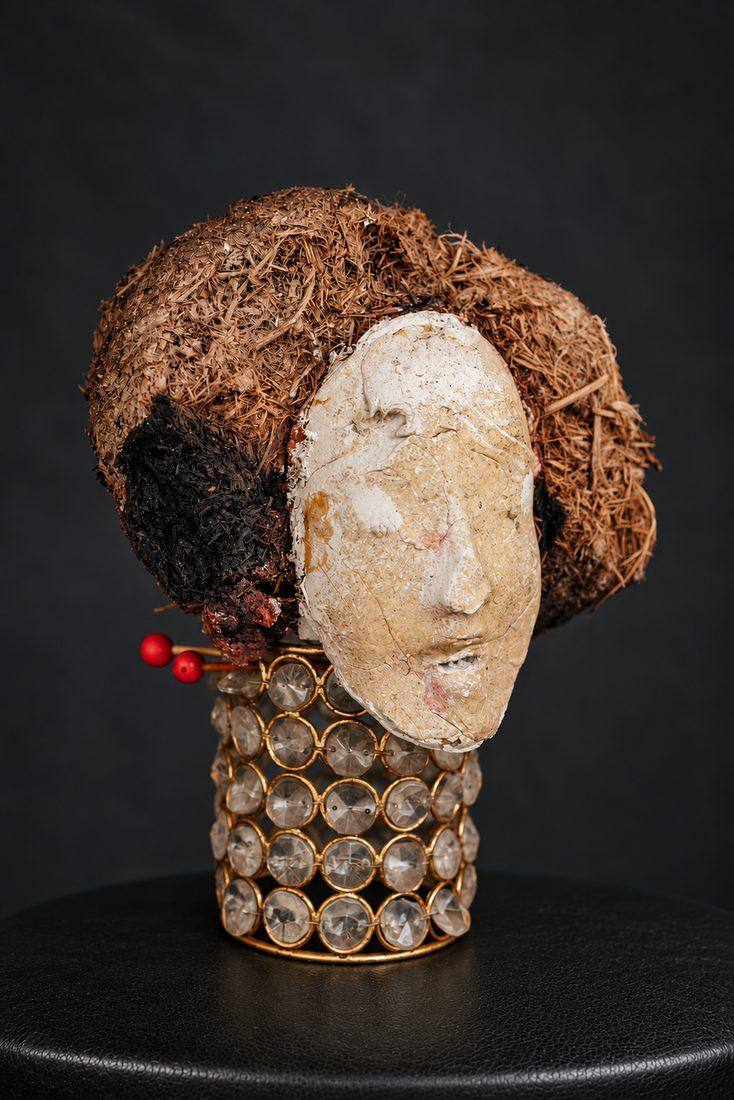 Pale moulded face set in dried brown fibre, two red-tipped pins, on a stand of cut-glass beads in gold rings, on black.