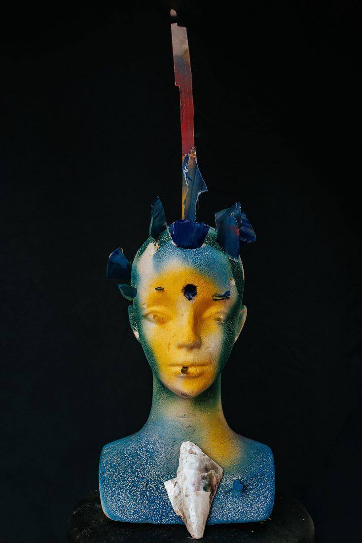 Bust sprayed yellow, blue and teal, blue glass shards in the head, a tall painted strip, a shell and a pinned blue heart on the chest, on black.