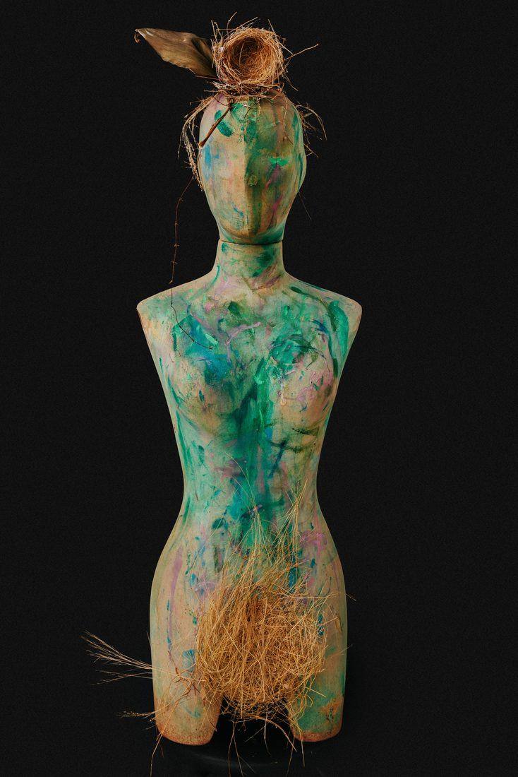 Dress form painted green, blue and pink, a bird's nest and a dried leaf on its head, dried grass at the hips, on black.