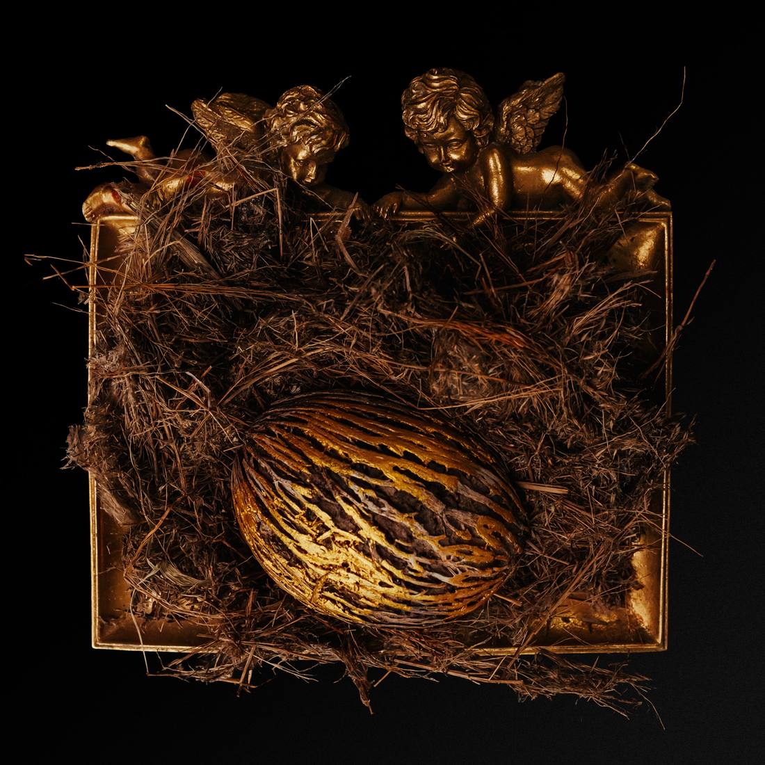 Seen from above on black: a gold frame filled with dried grass, a gold-streaked egg at the centre, two gilt cherubs at the top edge.