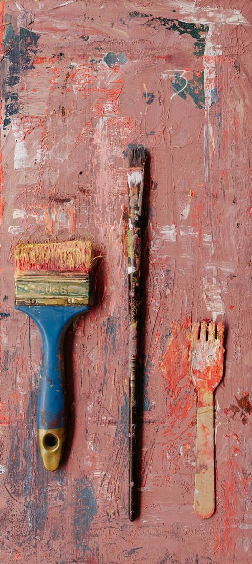 A wide brush with a blue handle, a long fine brush and a small wooden fork on a board thickly painted pink and red.