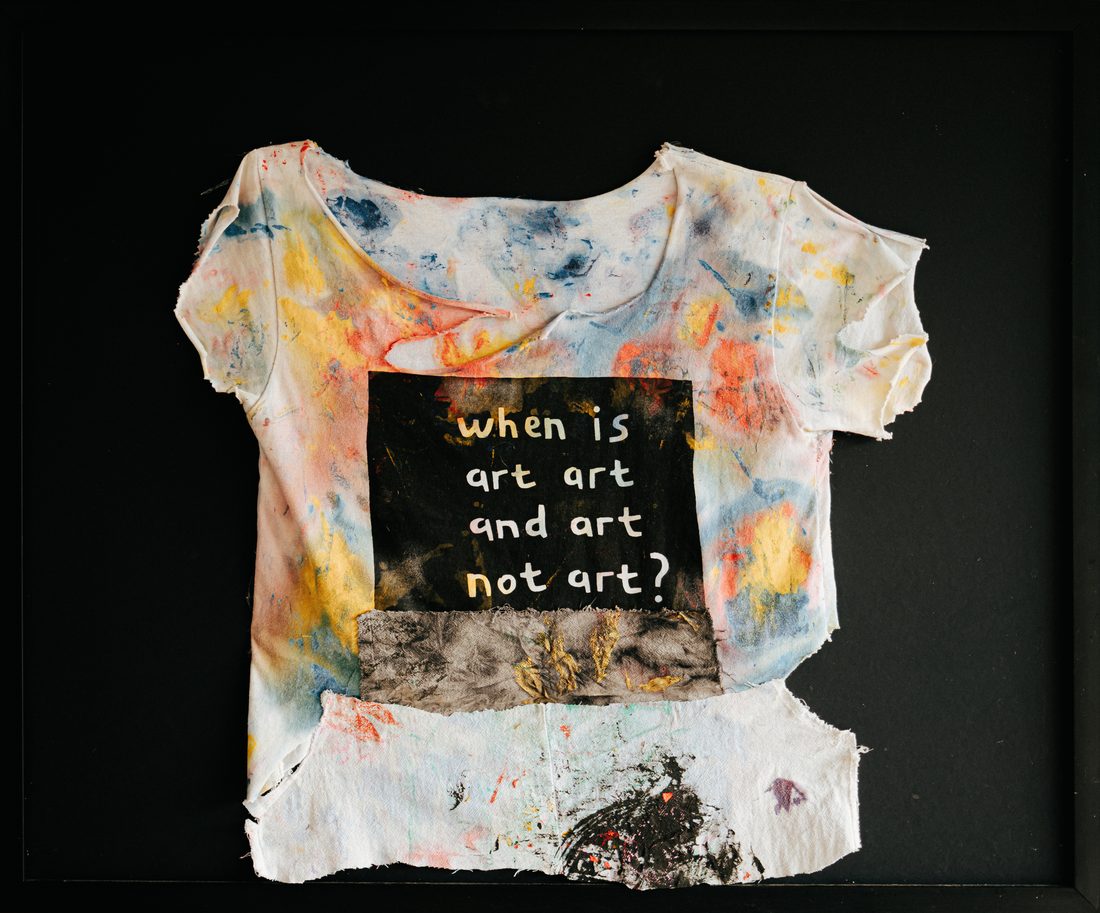 A cut-up white T-shirt splashed with colour, framed on black: a black panel lettered when is art art and art not art?, a grey strip with gold below.